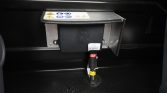 Electrical control module mounted on a metal bracket with warning labels and bundled cables passing through a floor grommet in a dark compartment.