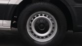 Close-up of a silver steel wheel with a black center cap and circular ventilation holes on a dark tire, mounted to a white vehicle panel visible nearby
