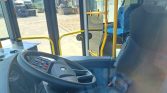 Interior view of a bus driver’s cockpit with a steering wheel, control panel, and blue patterned seats, outside yard visible through windows.