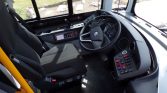 Interior of a bus cockpit featuring a steering wheel, gauges, and a row of control switches by the driver’s seat. The large windshield is visible ahead.