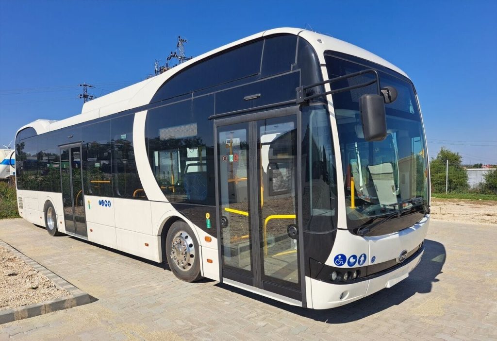 Front-side view of a white modern city bus with large curved windshield and open entry doors, parked on a brick lot under a blue sky.
