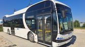 Front-side view of a white modern city bus with large curved windshield and open entry doors, parked on a brick lot under a blue sky.