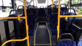 Interior of a city bus viewed from the rear aisle, blue patterned seats, yellow handrails, and a central aisle leading forward.