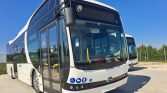 Front view of a white BYD city bus with accessibility icons on the lower front, parked on a paved lot with another bus in the background, and blue sky above.