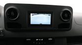 Infotainment screen showing rear-view camera feed in a car dashboard with round air vents on both sides and climate controls below (reverse view).