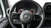 Mercedes-Benz steering wheel with integrated controls, inside a car cabin showing the instrument cluster in the background.