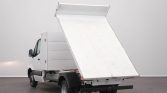 White delivery truck with its flatbed/dump bed raised at a steep angle in a studio setting behind it, showing a clean gray floor and light gray wall.