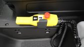 Yellow handheld control device with left and right arrow buttons and a red emergency stop button, connected by a coiled cord to vehicle equipment.