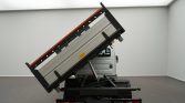 Rear view of a small IVECO Daily pickup/dump truck with its cargo bed raised at a steep angle inside a white studio setting