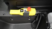 Yellow handheld control with left/right arrow buttons and red emergency stop, attached by a coiled cord in a vehicle interior (likely for a lift or accessibility device).