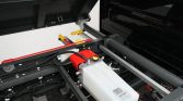 Industrial hydraulic system under a vehicle chassis, featuring a white reservoir tank, red covers, and yellow levers with a gray support beam nearby.