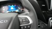 Close-up of a car steering wheel with cruise control buttons on the right and a blurred digital dashboard in the background.