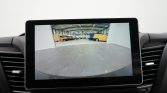 Car backup camera display showing a warehouse with yellow vans; Dutch message reads 'Let op de omgeving' (Watch surroundings).