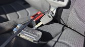 Close-up of a car interior showing a seat belt buckle area with a small black device labeled VEGO attached and a key nearby on black fabric upholstery.