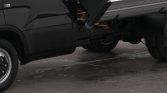 Black pickup truck with a raised metal dump bed visible on the right, wheel on the left, parked on a concrete surface.