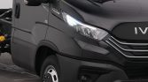 Close-up of the front-right side of a dark gray Iveco cargo van, with a bright headlight and visible grille and wheel.