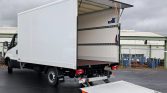 White box truck with a lowered rear loading ramp (tail lift) exposing an empty cargo area ready for loading near an industrial site.
