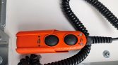Orange handheld control pendant with two large black pushbuttons for lifting and lowering, connected to a coiled cable on a white surface.
