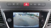 Car dashboard touchscreen showing a rear-view camera feed with a parking lot and construction equipment behind the vehicle