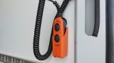 Orange control pendant with two circular black buttons and a coiled cord, mounted on a white industrial panel.