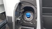 Open fuel-filler door on the side of a white van, showing a blue fuel cap and surrounding fuel-entry area.