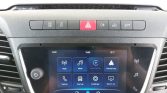 Car dashboard with a touchscreen showing infotainment apps (Radio, Media, Apple CarPlay, Android Auto) and a row of control buttons above it, including a red hazard button in the center.