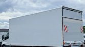 White box truck trailer viewed from the side with a cab visible on the left, parked in a lot under a cloudy sky.