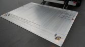 Aluminum loading ramp attached to a truck, viewed from above, resting on asphalt with small orange lights on its edges.