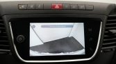 Dashboard touchscreen showing the rearview camera feed for parking, with guidance lines and the Dutch message 'Controleer de hele omgeving' on screen.