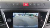 Car infotainment screen showing a rear-view camera feed of a parking area, with 'Controleer de hele omgeving' warning text in the image.