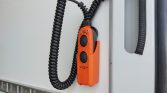 Orange two-button control pendant with a coiled cord, mounted inside a vehicle, used to operate equipment remotely.