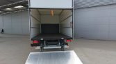 Rear of an open box truck with a metal loading ramp extended to the ground in a paved lot; interior empty and ready for cargo, orange strap hanging from ceiling.