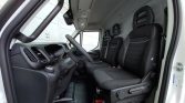 Front cabin of a white commercial van showing the dashboard, steering wheel, and three black upholstered seats with headrests, viewed from the open driver's door