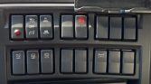 Close-up of a vehicle's dashboard control panel: row of black rectangular switches, including a red hazard-light button in the center and several OFF/AUTO labels.