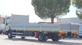 Flatbed truck parked in an industrial yard with a large tree in the background, white and gray cab.