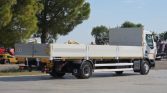 Side view of a white flatbed truck with high side rails, parked in a yard with trees and yellow machinery in the background.