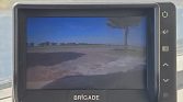 In-car display labeled BRIGADE showing a rural landscape with a tree, blue sky, and gravel road in the foreground