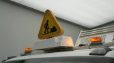 Yellow triangular roadwork sign with a worker icon mounted on top of a vehicle, orange warning lights illuminated start/stop.