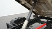 Hydraulic piston supporting a tilted trailer bed, with a metal frame and a red safety bar in view.