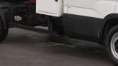 Side view of a white truck with a black lower panel, showing its undercarriage and a circular ground hatch marked with a yellow X tape.