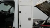 White utility truck with a side storage cabinet between the cab and the dump bed; hydraulic dump mechanism visible on the right.