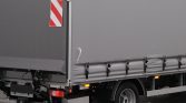 Grey cargo trailer with a red-and-white diagonal hazard panel on the rear door and a taut grey side tarp, wheel visible in the foreground.