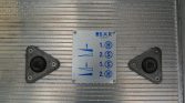 Metal control plate with blue arrows and H/S labels (lift indicators) mounted on a ribbed metal surface, flanked by two triangular black caps.