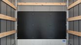 Interior of a cargo trailer: a large black panel mounted on the rear wall, with wooden rail supports along the sides and a light gray base at the bottom edge.