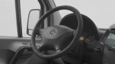 Interior of a vehicle cab showing a steering wheel with a Mercedes logo, dashboard, and side mirror visible through the window.