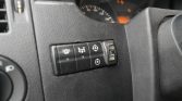 Close-up of a row of black vehicle controls on the dashboard left of the steering wheel, with light and drivetrain icons and a blurred instrument cluster in the background.