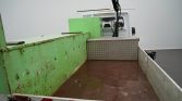 Flatbed truck with a worn green side panel, rusty brown bed, and a diamond-plate toolbox at the front of the bed. Truck cab visible in the background, with a crane arm above.