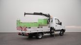 White utility truck with a mounted crane on a flatbed, green equipment box, and red-and-white striped rear—stabilizer leg extended on a dark workshop floor.