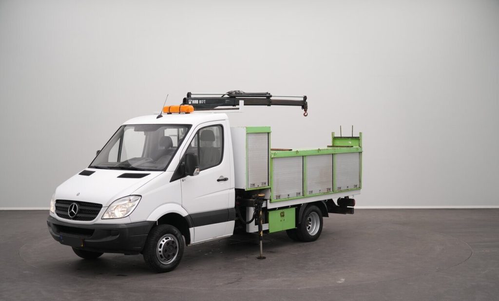 White Mercedes-Benz utility truck with a mounted crane on top and green cargo bed, parked indoors on a gray backdrop.
