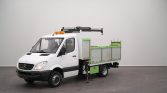 White Mercedes-Benz utility truck with a mounted crane on top and green cargo bed, parked indoors on a gray backdrop.